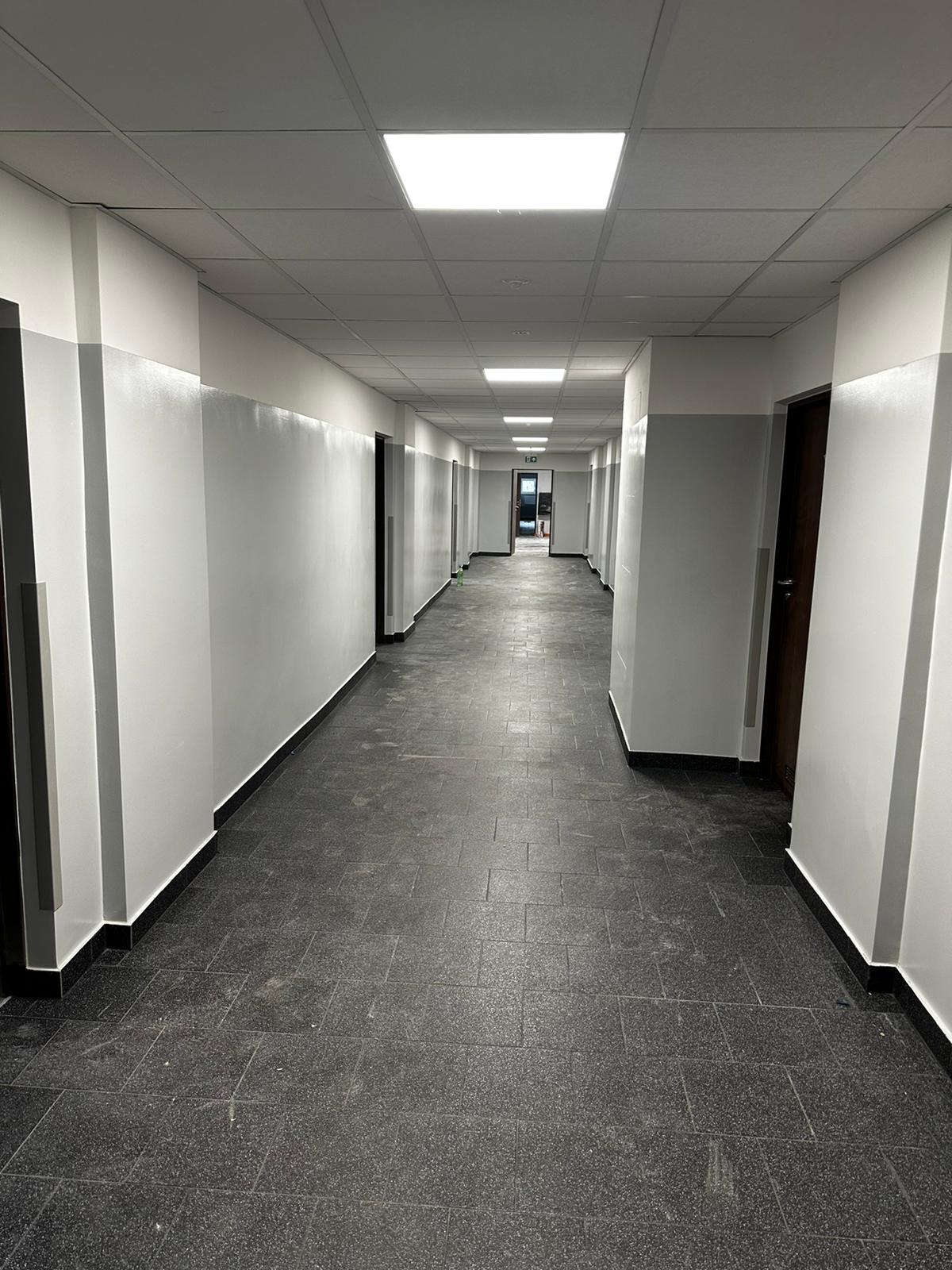Długi korytarz w budynku biurowym z szarą podłogą z płytek, białymi ścianami z szarym pasem i szeregiem drzwi po obu stronach, oświetlony panelami sufitowymi.