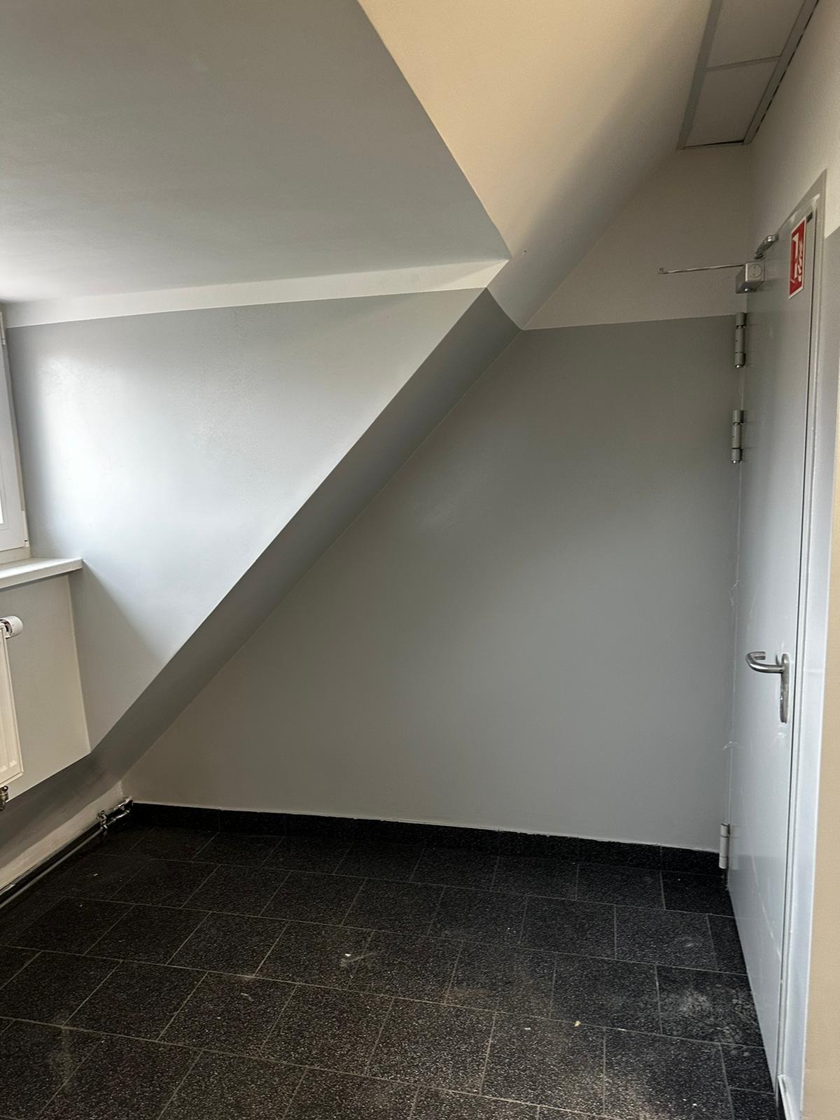 Pomieszczenie po remoncie: świeżo pomalowane szare ściany z nietypowym skosem sufitu, ciemna, błyszcząca podłoga z płytek, białe drzwi z metalową klamką.