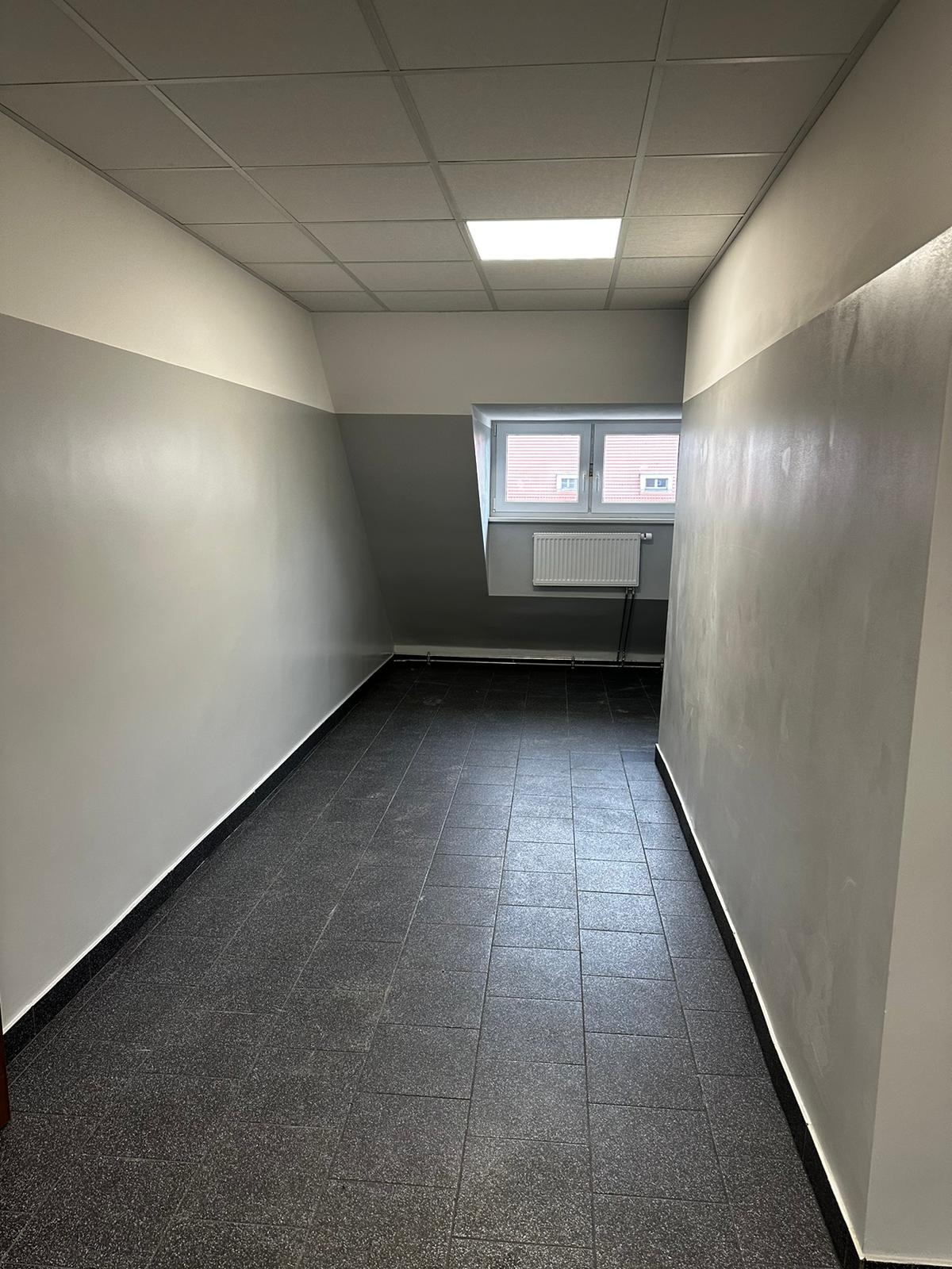 Wnętrze korytarza po remoncie: szare ściany z białym pasem u góry, ciemna, kwadratowa podłoga z płytek, sufit podwieszany z lampą, widoczne okno z grzejnikiem.