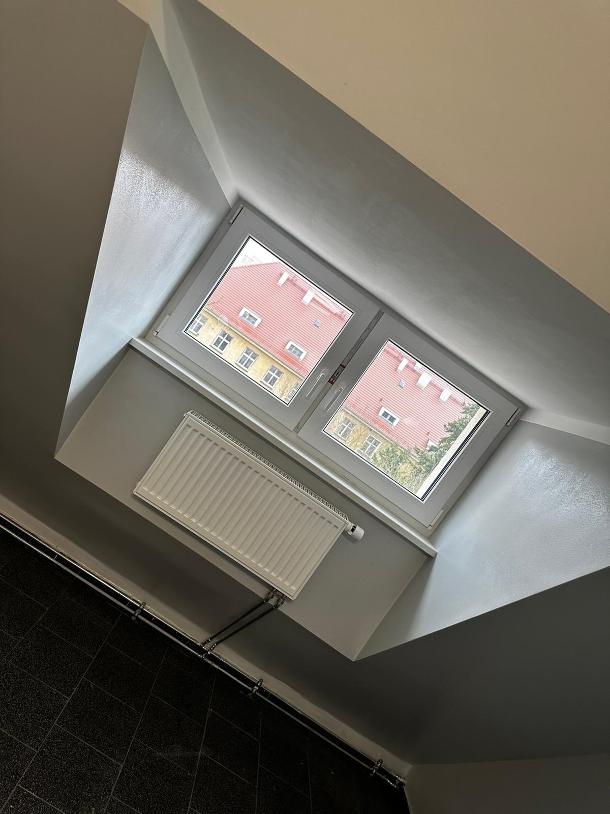 Poddasze po szpachlowaniu i malowaniu na szaro, z widocznym oknem dachowym, grzejnikiem i fragmentem czarnej podłogi.