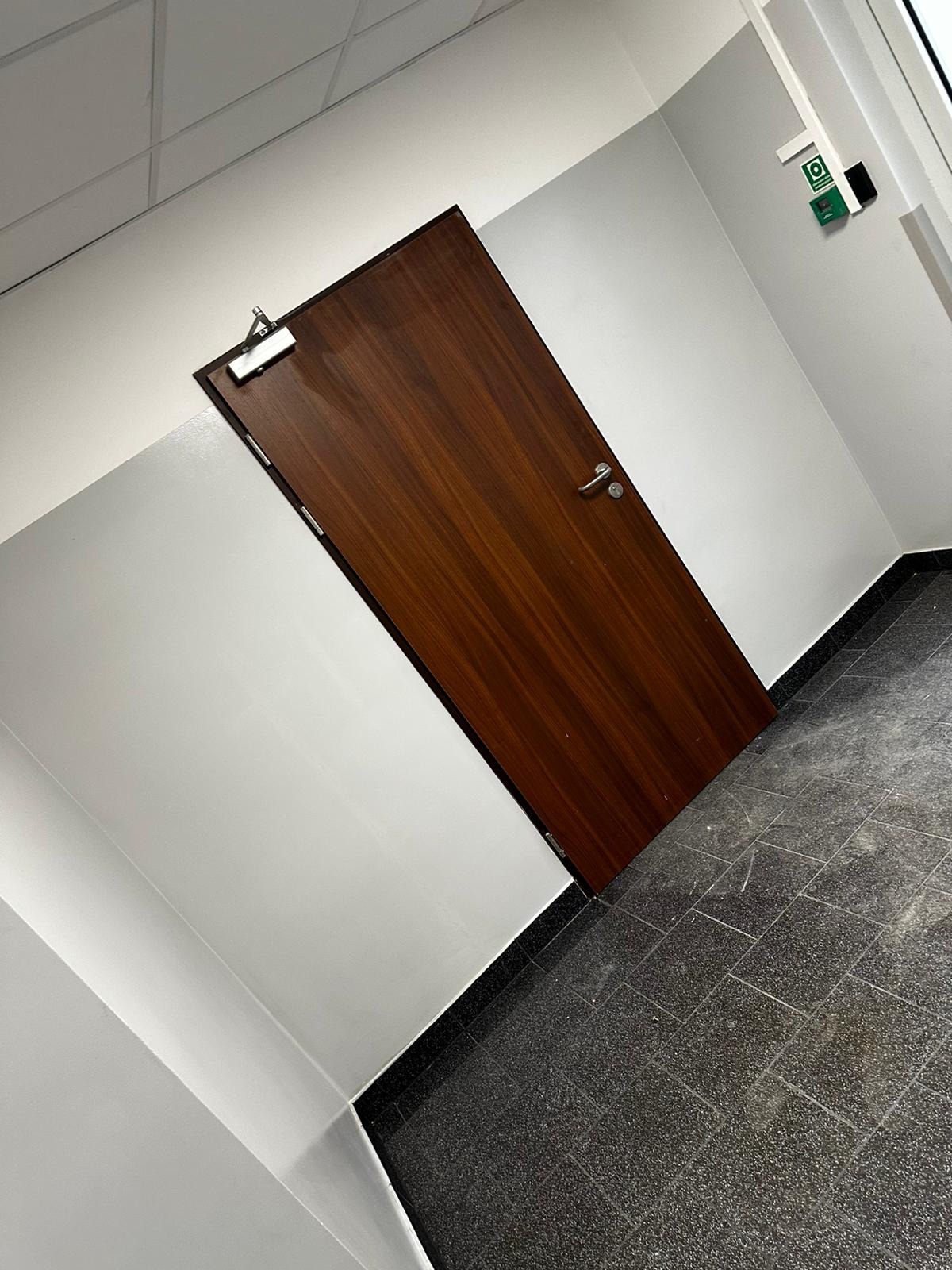 Ciemnobrązowe drzwi z chromowanymi okuciami w białym pomieszczeniu z szarą podłogą z płytek. Widoczny samozamykacz na górze drzwi i czujnik bezpieczeństwa na ścianie.