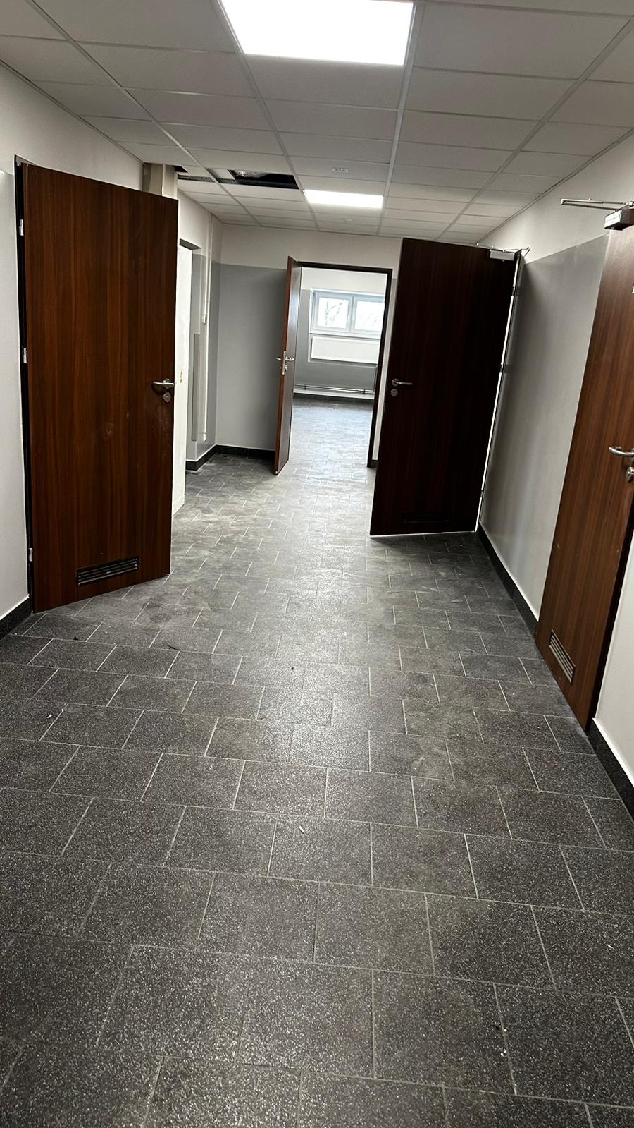 Korytarz biurowy z szarą terakotą na podłodze i drewnianymi drzwiami, widoczne niedociągnięcia w suficie podwieszanym.