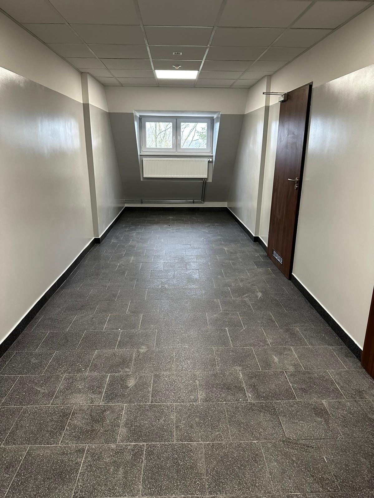 Wnętrze korytarza z szarą podłogą z płytek, biało-szare ściany z oknem na końcu i brązowe drzwi po prawej stronie.