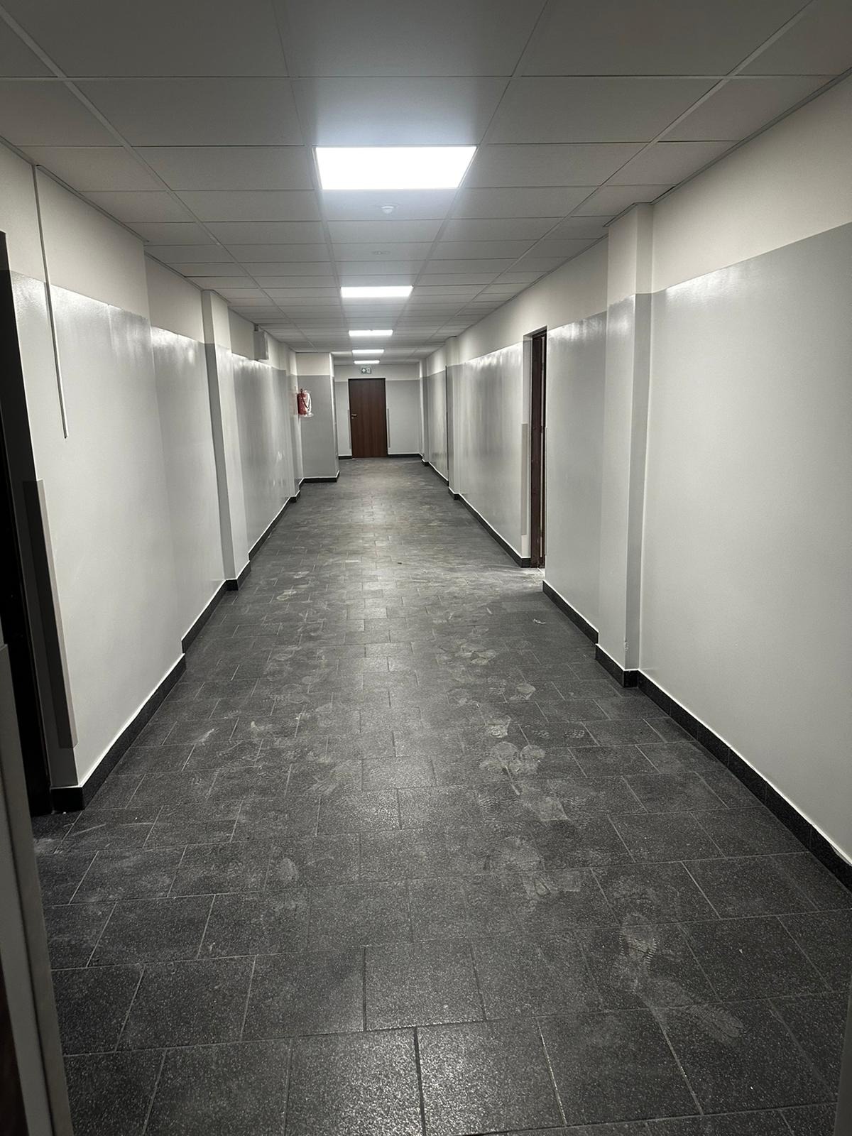 Długi korytarz biurowy z szarą podłogą z płytek i świeżo pomalowanymi na biało ścianami z szarym pasem, widoczne drzwi i oświetlenie sufitowe.