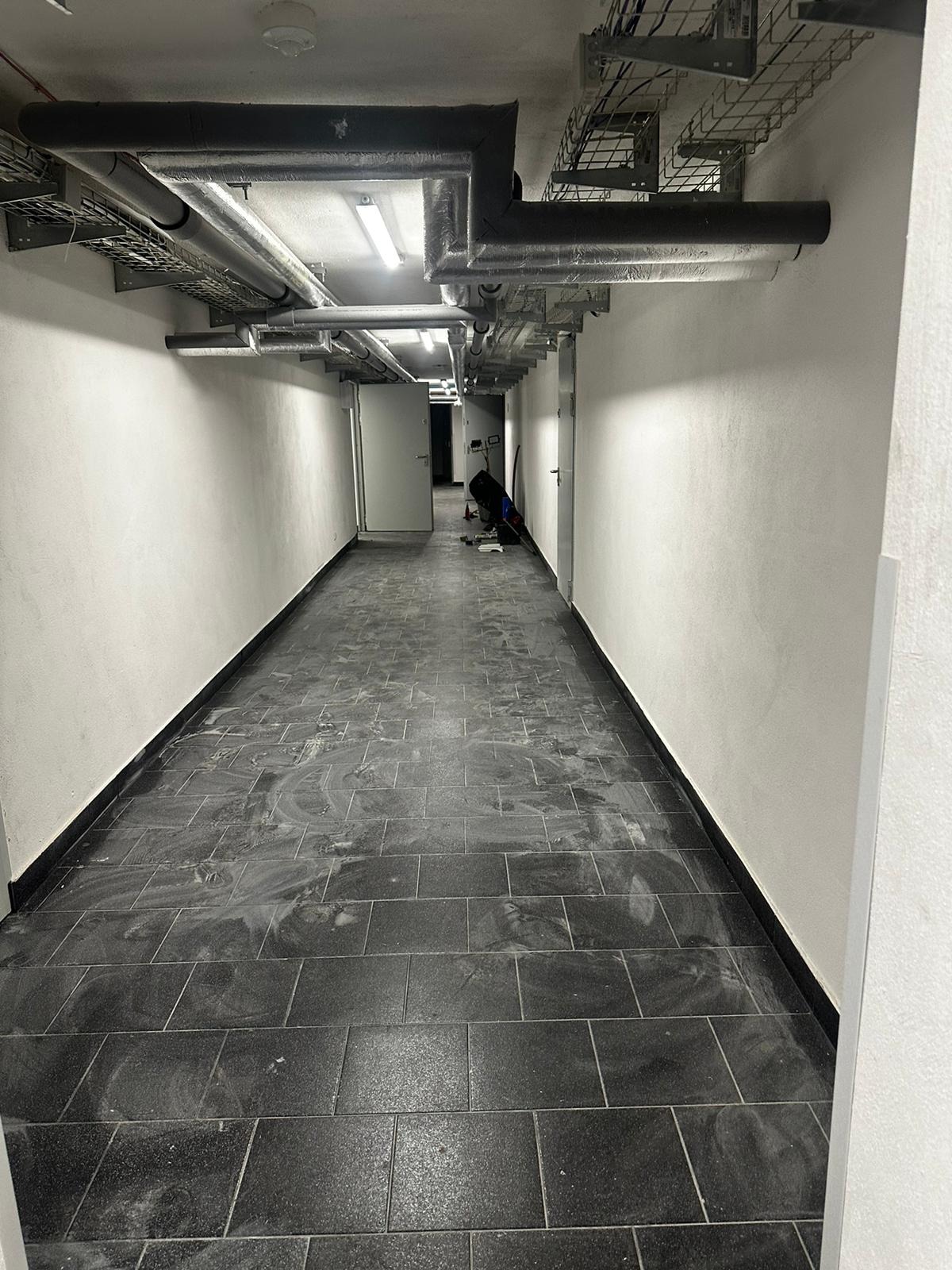 Korytarz z ciemną, prostokątną posadzką w trakcie czyszczenia, widoczne rury pod sufitem i osoba pracująca w oddali.
