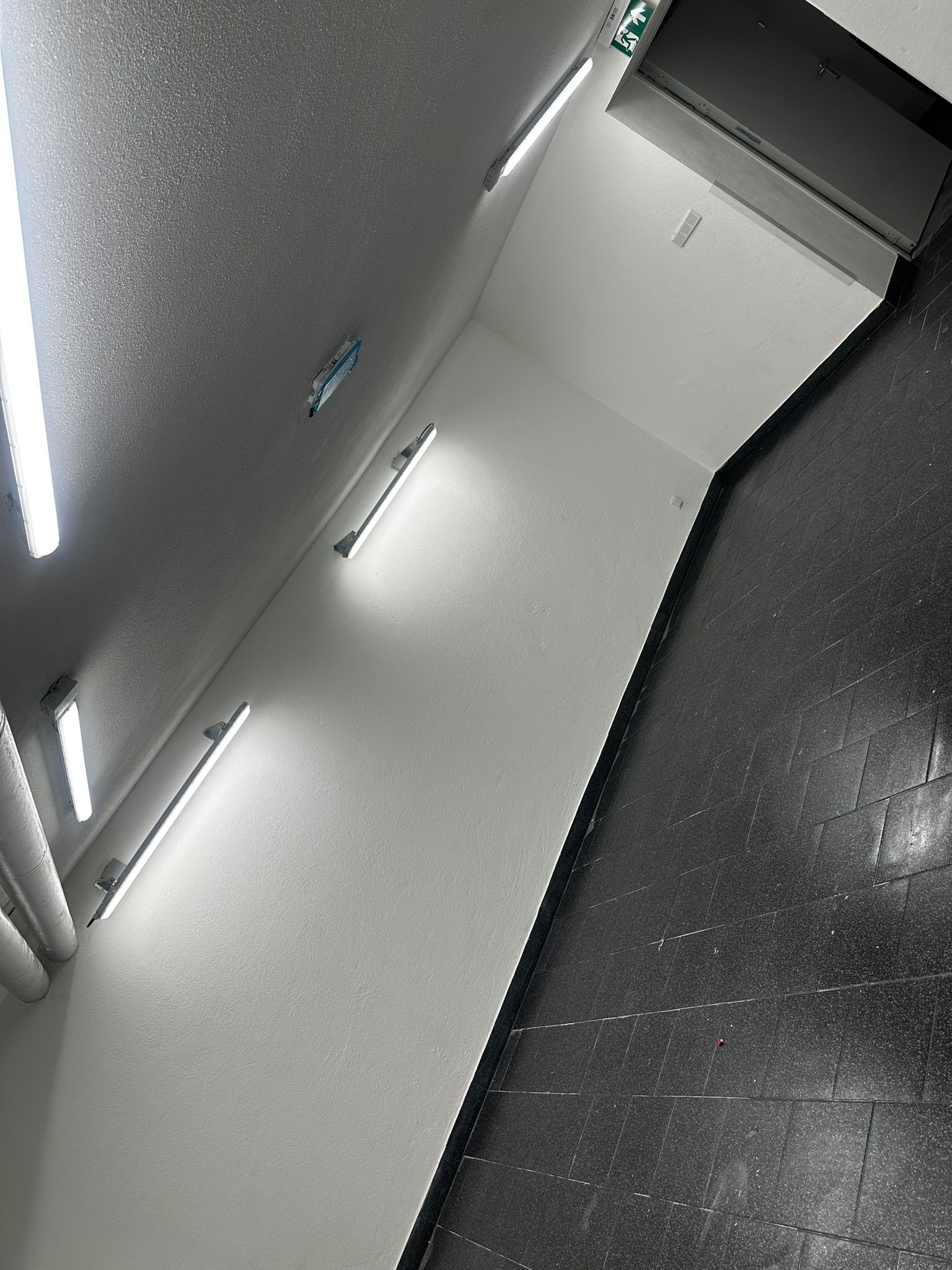 Wnętrze korytarza po remoncie: świeżo pomalowane, białe ściany z rzędami lamp fluorescencyjnych, czarna, błyszcząca podłoga z płytek, widoczne rury przy ścianie, częściowo zabezpieczone miejsce po...