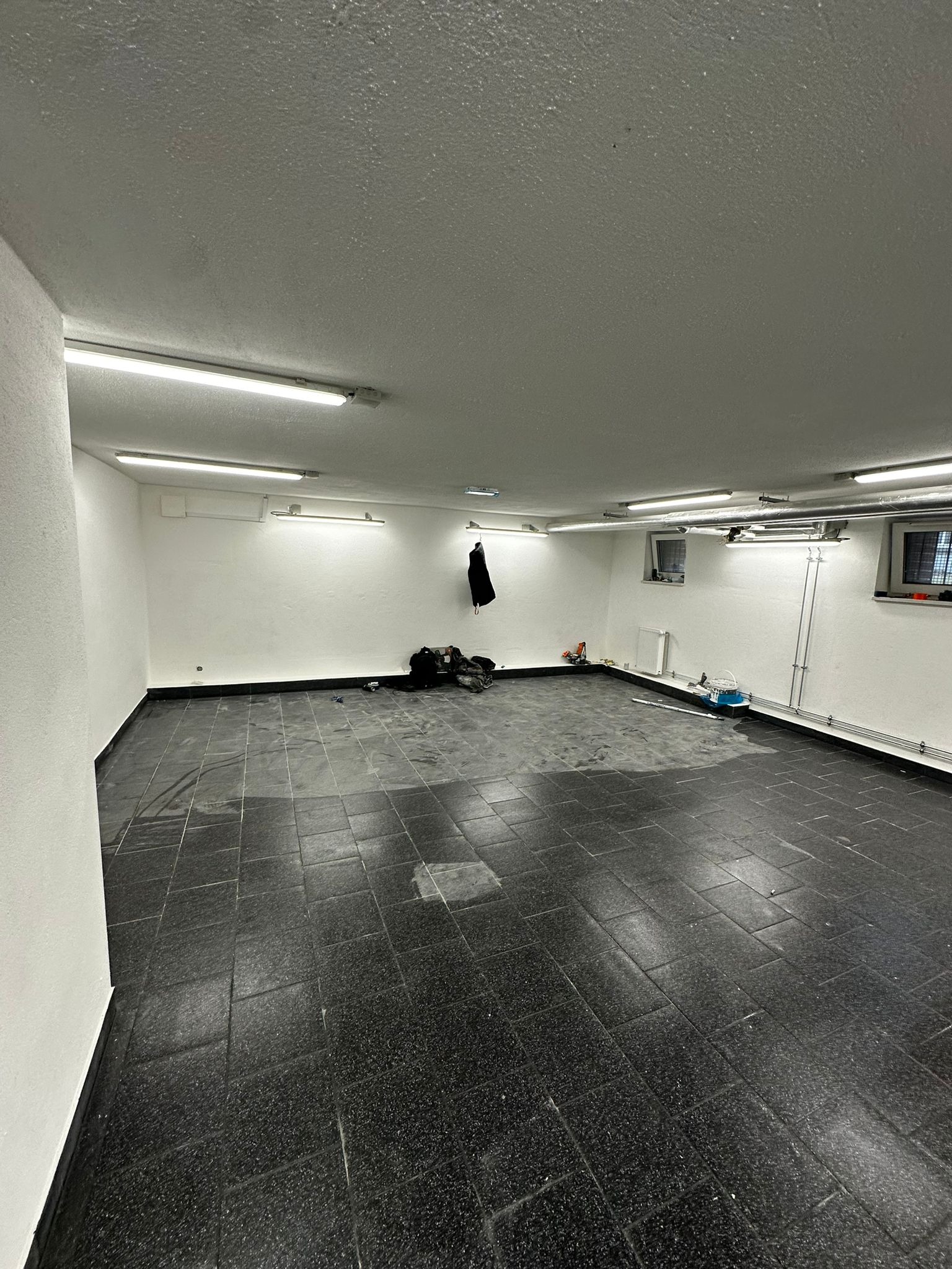 Pusta przestrzeń z czarną, kwadratową podłogą z płytek, białe ściany, oświetlenie jarzeniowe, okna w tle i narzędzia w rogu, przygotowana do remontu.