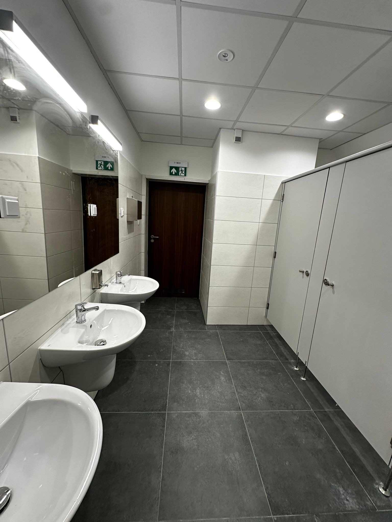 Wnętrze publicznej toalety z trzema umywalkami, lustrem, płytkami na ścianach i podłodze oraz kabinami WC z białymi drzwiami.