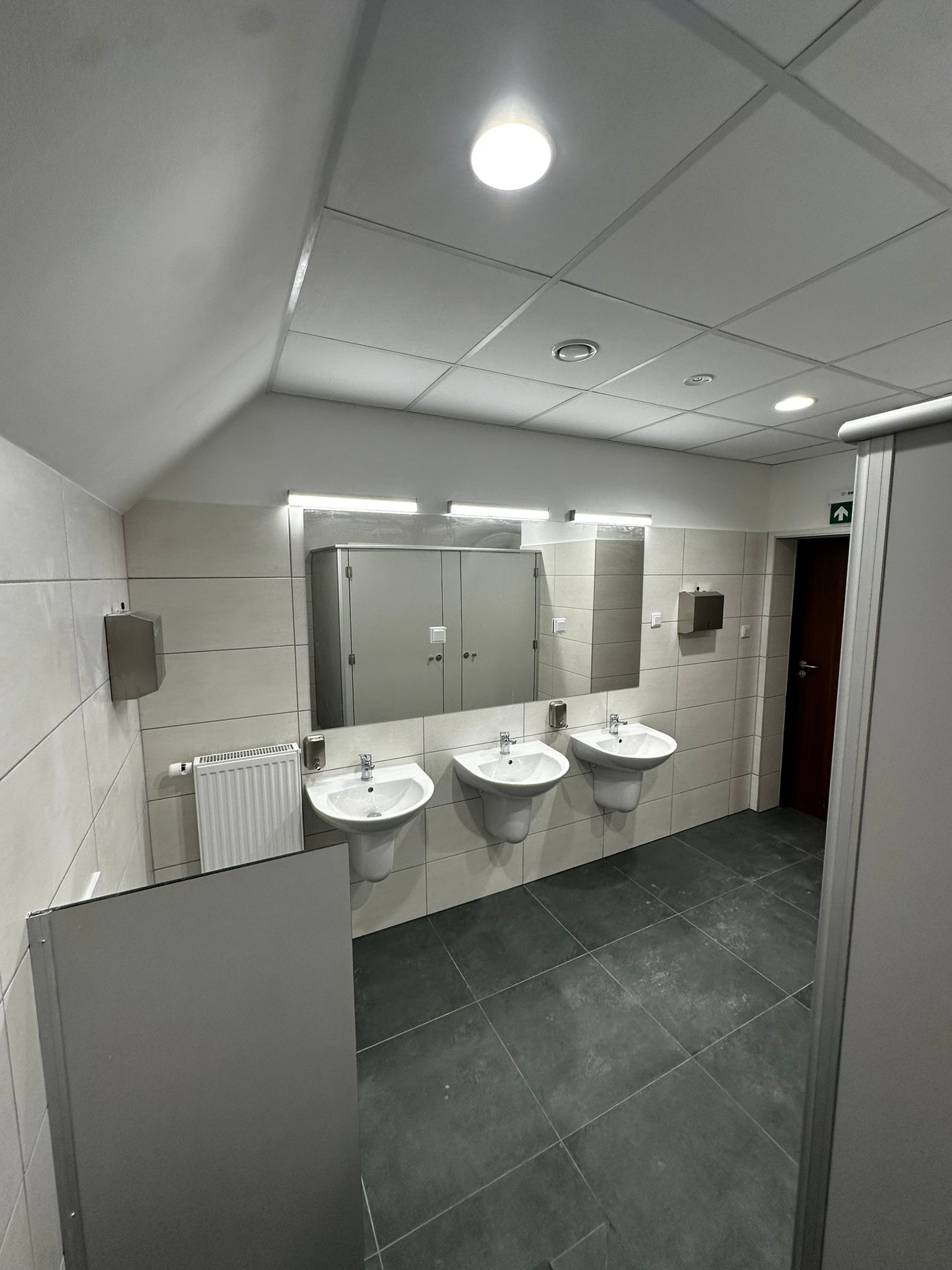 Wnętrze publicznej toalety z trzema umywalkami na postumentach, dużym lustrem z oświetleniem LED, szarymi płytkami ściennymi i podłogowymi, oraz kabinami WC z drzwiami.