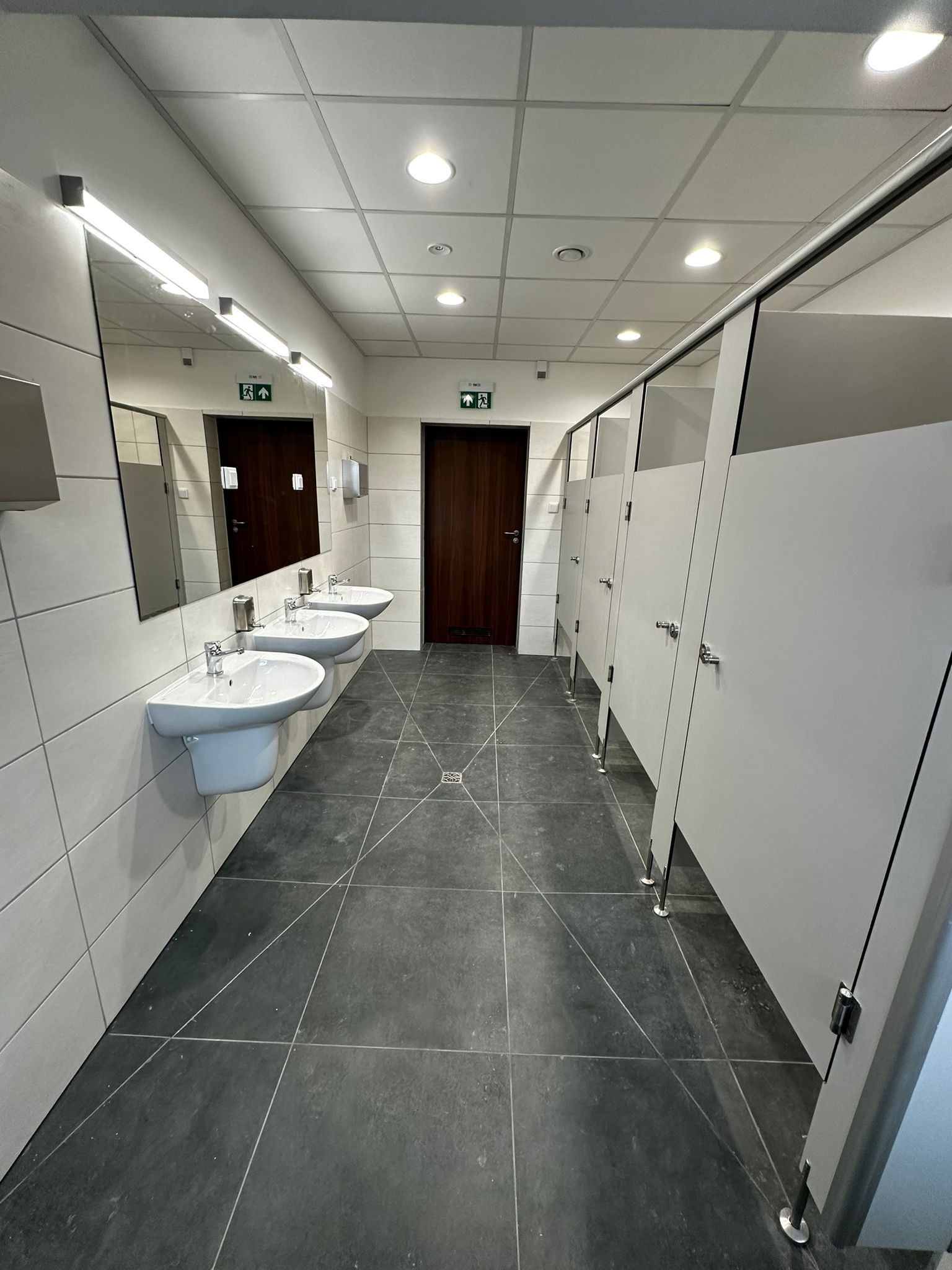 Wnętrze publicznej toalety z trzema umywalkami, dużym lustrem z oświetleniem, kabinami WC z szarymi drzwiami i ciemną podłogą z płytek. Nad drzwiami widoczne znaki ewakuacyjne.
