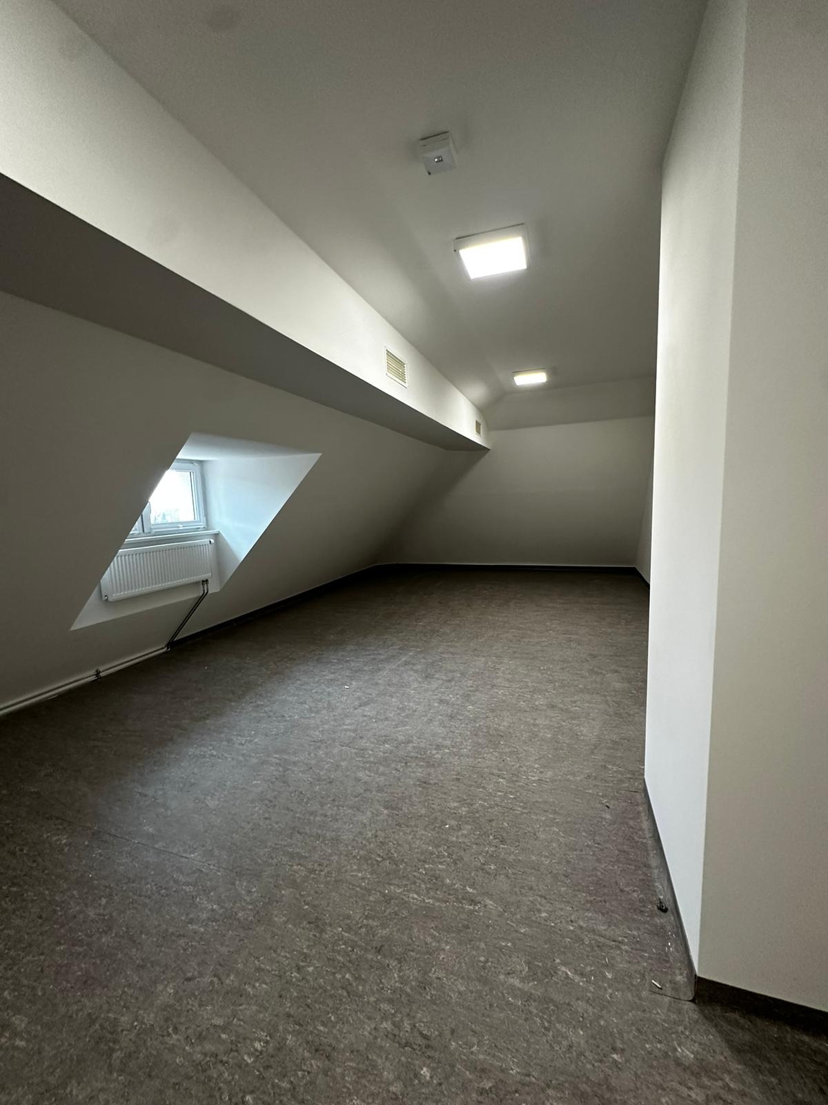 Puste poddasze ze skośnym oknem, szarym panelem podłogowym i białymi ścianami, przygotowane do dalszego zagospodarowania.