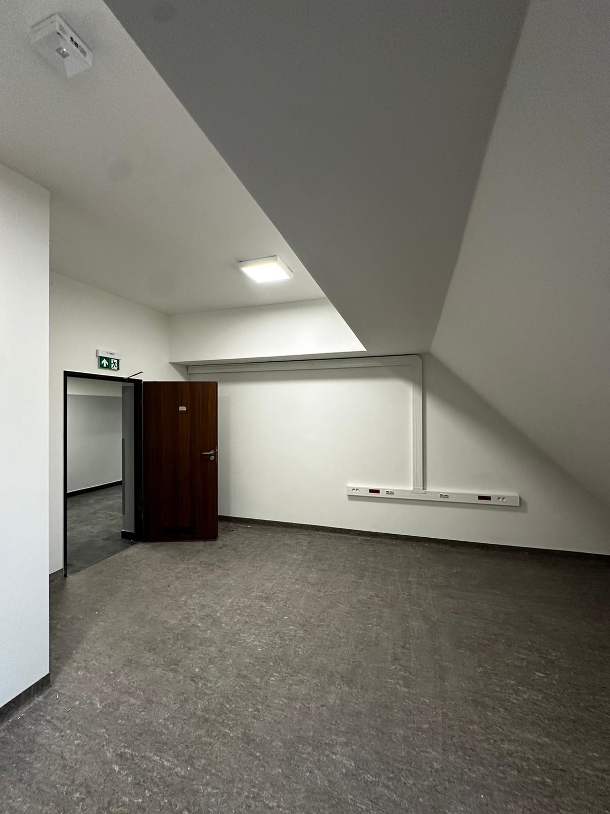 Puste, szare pomieszczenie biurowe z otwartymi drzwiami, białymi ścianami, szarą podłogą i skośnym sufitem, widoczne instalacje elektryczne.