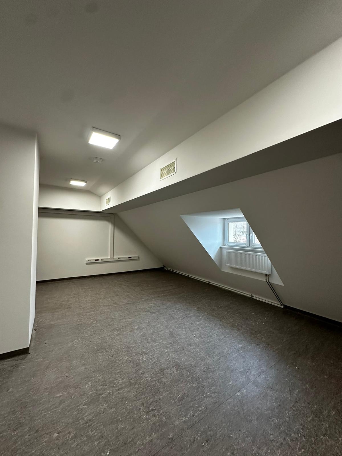 Puste poddasze z szarą wykładziną podłogową, skośnym sufitem, oknem w kształcie trapezu i białymi ścianami, przygotowane do dalszych prac wykończeniowych.