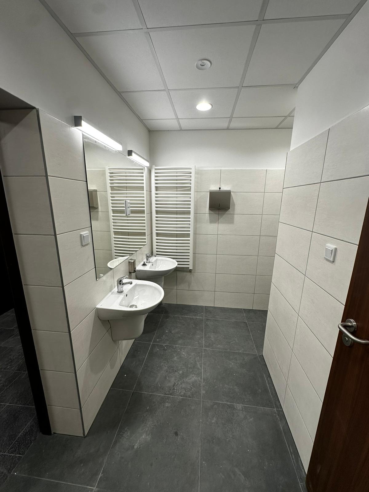 Wykończona łazienka publiczna z dwiema umywalkami ceramicznymi na postumentach, grzejnikami drabinkowymi i szarą podłogą z płytek. Widoczne płytki ścienne w odcieniach bieli i szarości...