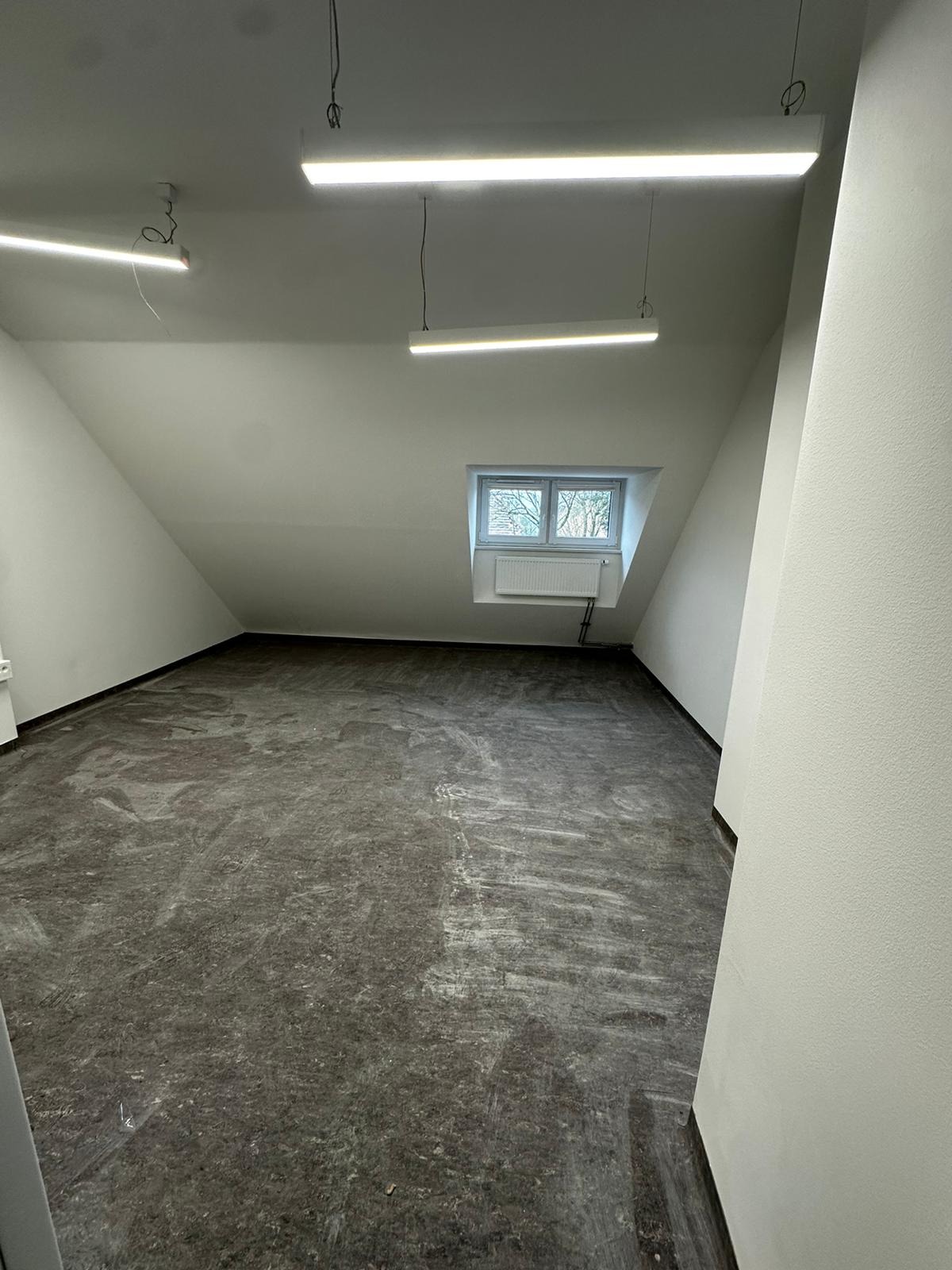Pokój na poddaszu w trakcie prac wykończeniowych, widoczne przygotowane podłoże pod panele, białe ściany, okno w szczycie dachu i liniowe oświetlenie LED.