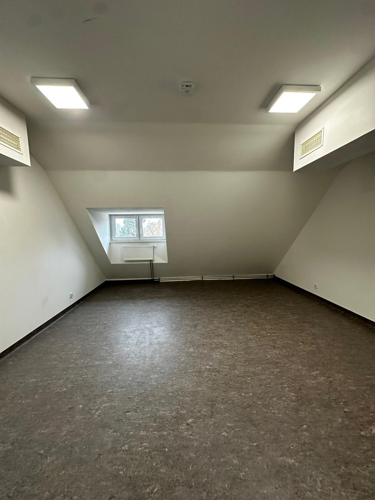 Puste poddasze z brązową podłogą, oknem w skosie dachu, białymi ścianami i dwoma kwadratowymi lampami sufitowymi.