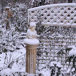 Prestiż - Zimowy ogród z rzeźbą anioła na kolumnie, białą kratownicą i roślinami w koszach, wszystko pokryte śniegiem.