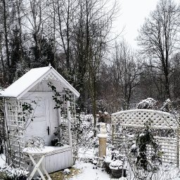 Prestiż - Zimowy ogród z białym domkiem narzędziowym i pergolą pokrytymi śniegiem, dekoracyjne rośliny w donicach również pod śniegiem, drzewa bez liści w tle.