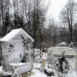 Zimowy ogród z białym domkiem narzędziowym i pergolą pokrytymi śniegiem, dekoracyjne rośliny w donicach również pod śniegiem, drzewa bez liści w tle.