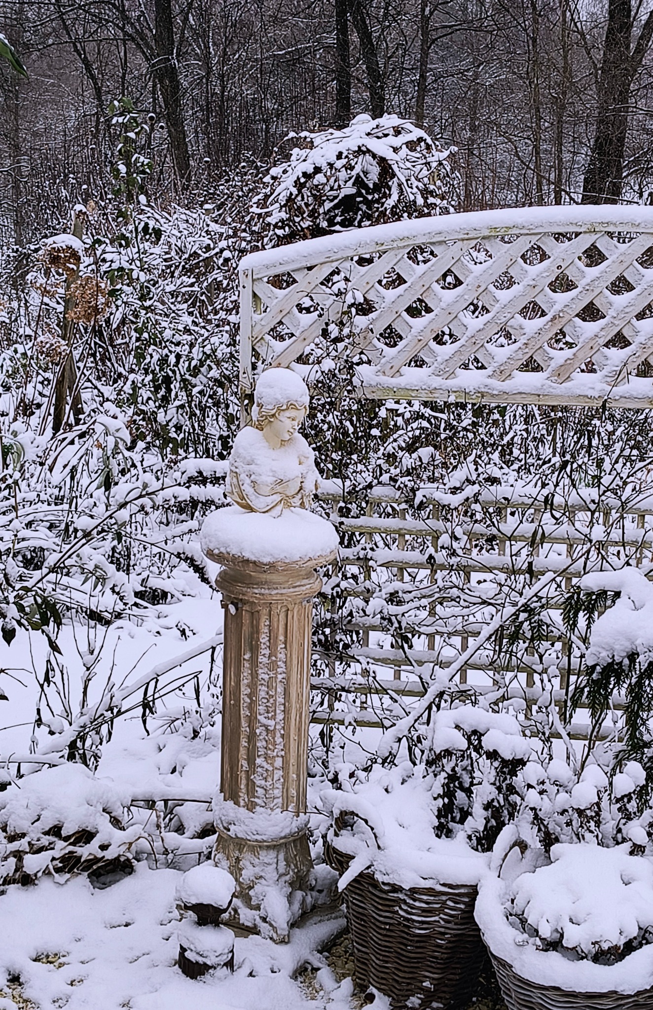 Zimowy ogród z rzeźbą anioła na kolumnie, białą kratownicą i roślinami w koszach, wszystko pokryte śniegiem.