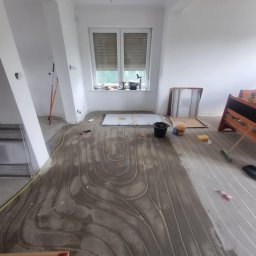 MTP Damian Lewandowski - Montaż ogrzewania podłogowego w pokoju, widoczne rurki zatopione w wylewce, narzędzia i materiały budowlane rozłożone na podłodze, okno z roletami w tle.