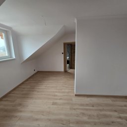 MTP Damian Lewandowski - Poddasze po remoncie: świeżo pomalowane białe ściany, jasna drewniana podłoga, okno z widokiem na zewnątrz, widoczne listwy przypodłogowe i otwarte drzwi do kolejnego pomieszczenia.