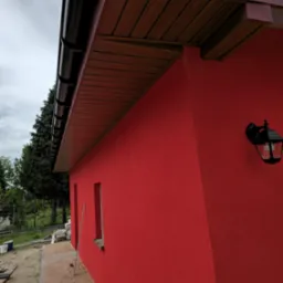 Czerwona elewacja domu z czarną lampą zewnętrzną i brązowym podbitką dachową. Widoczny fragment chodnika z kostki brukowej i zielone drzewa w tle.