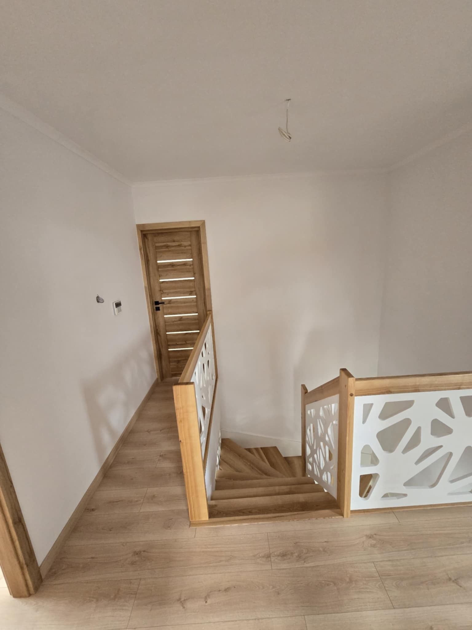 Wnętrze domu po remoncie: drewniane schody z białą, ażurową balustradą, drewniana podłoga i drzwi, białe ściany, wiszący kabel od lampy.