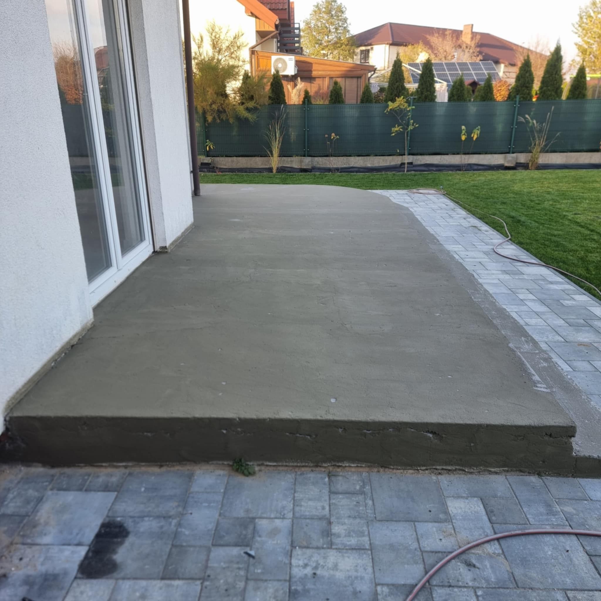 Wylewka betonowa tarasu w trakcie realizacji, widoczne połączenie z kostką brukową i budynkiem, ogród w tle.