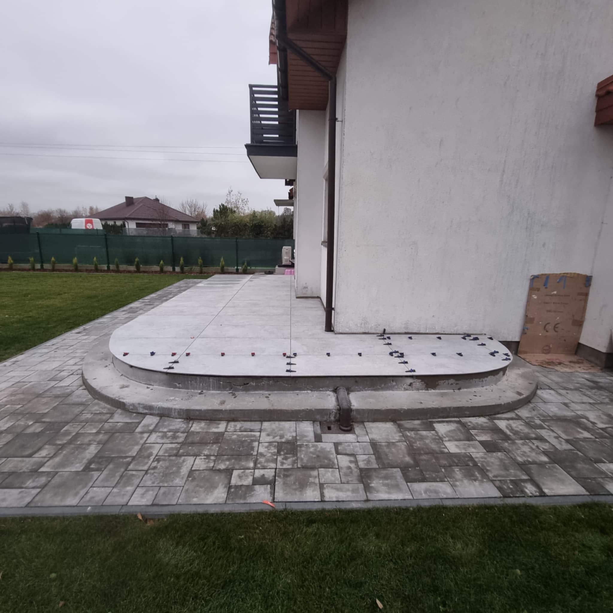 Układanie płyt betonowych na tarasie z użyciem poziomujących klipsów i klinów, widoczne obrzeża z kostki brukowej i trawnik, etap prac wykończeniowych