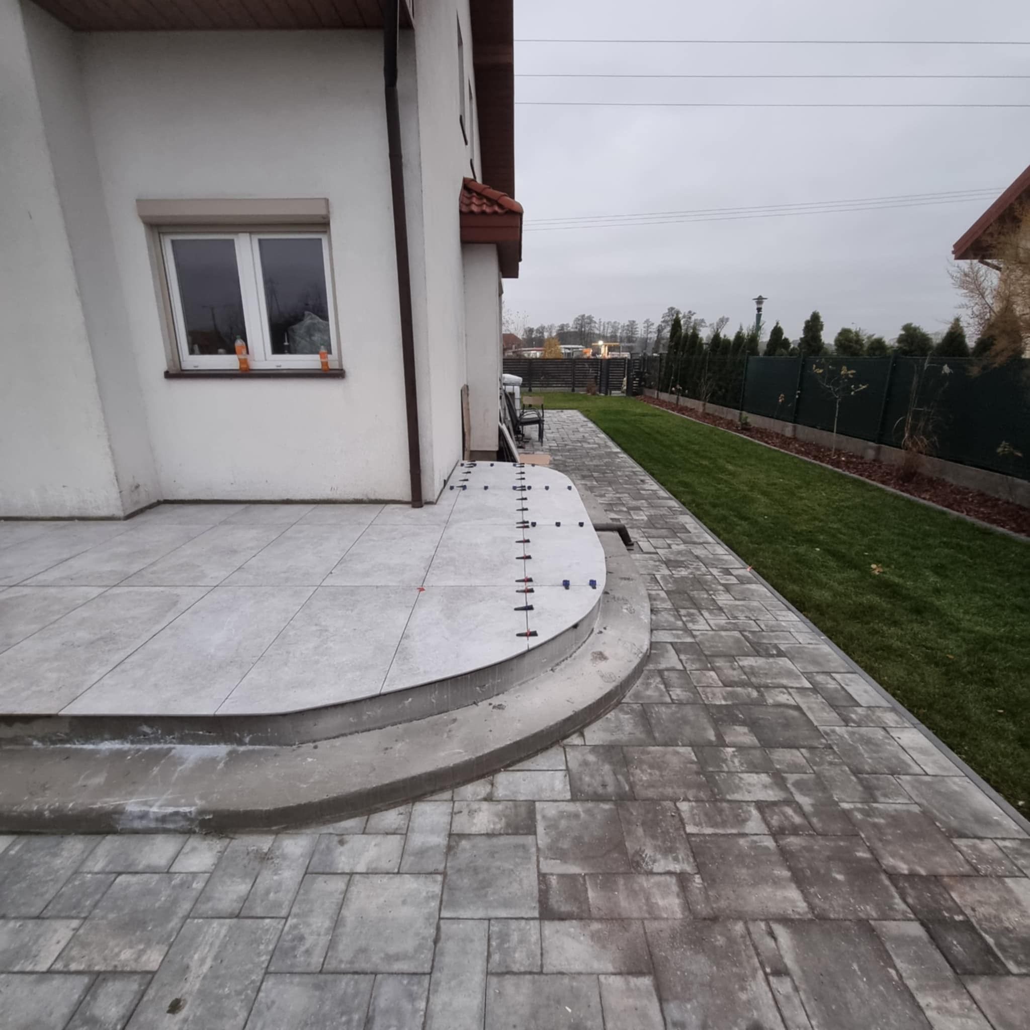 Układanie jasnoszarych płytek na tarasie przed domem, widoczne elementy poziomujące, trawnik i ogrodzenie w tle.