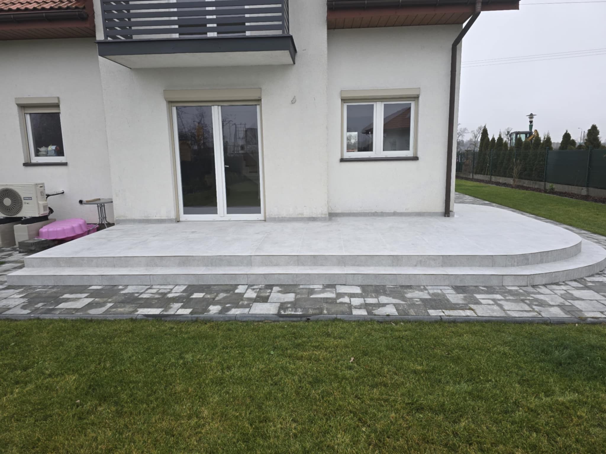 Wykończony taras z szarych płytek ceramicznych z zaokrąglonymi schodami, przylegający do białej elewacji domu z balkonem i szarą kostką brukową oraz zielonym trawnikiem.
