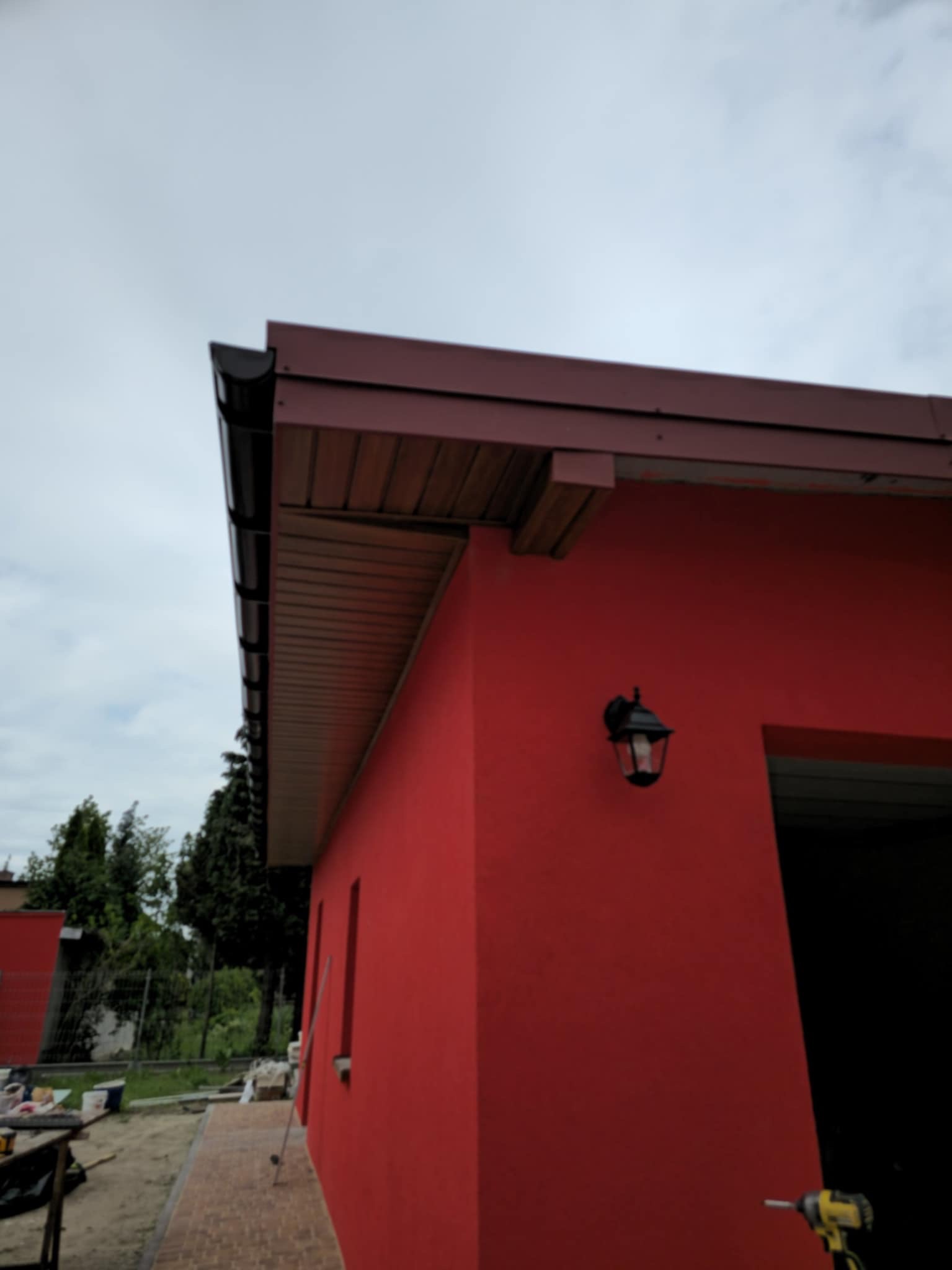 Czerwona elewacja budynku z brązowym dachem i czarnymi rynnami. Widoczna lampa zewnętrzna i narzędzia, sugerujące prace wykończeniowe.
