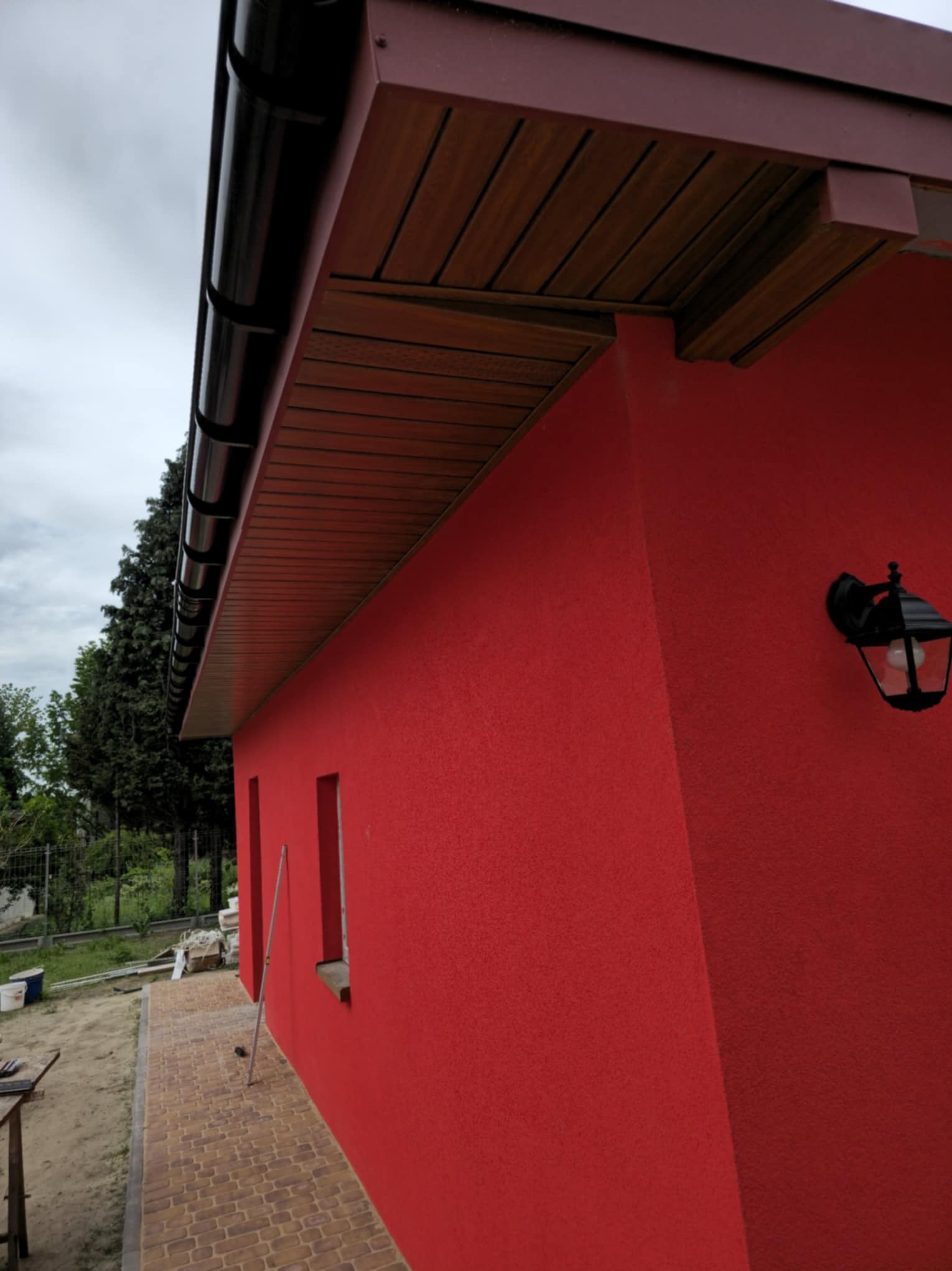 Czerwona elewacja domu z czarną lampą zewnętrzną i brązowym podbitką dachową. Widoczny fragment chodnika z kostki brukowej i zielone drzewa w tle.