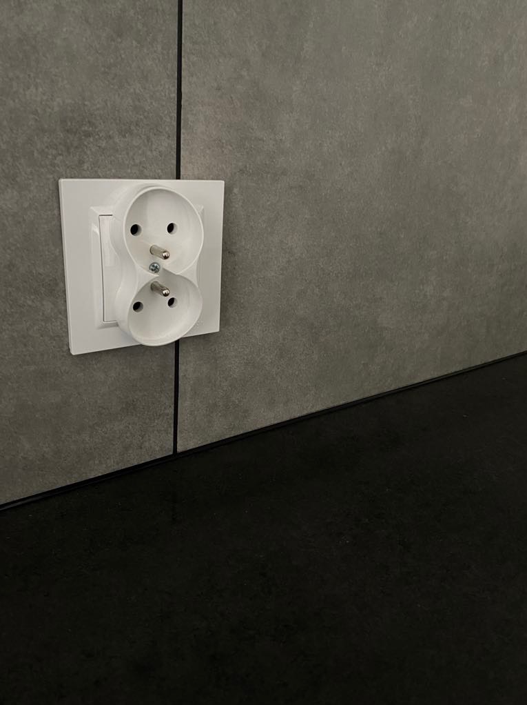 Podwójne, białe gniazdko elektryczne zamontowane na szarej, strukturalnej ścianie nad czarnym blatem. Widok z bliska, neutralne oświetlenie.