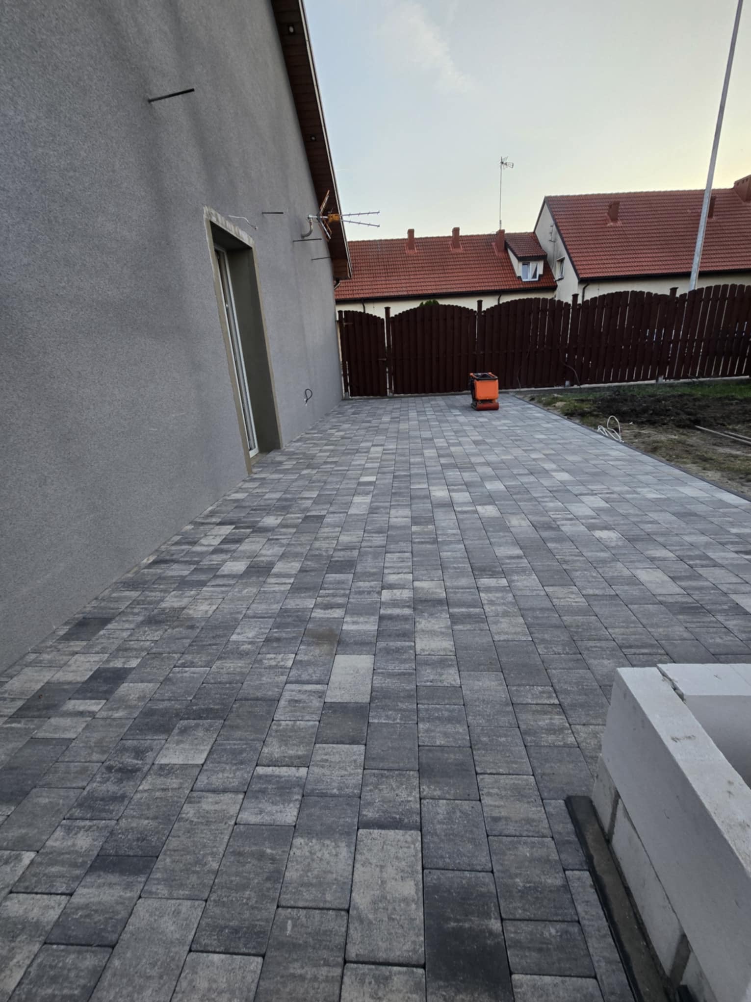 Ułożona kostka brukowa na posesji, szara kolorystyka. W tle ogrodzenie z brązowych desek i budynki mieszkalne.  Widok na świeżo wykonany podjazd.