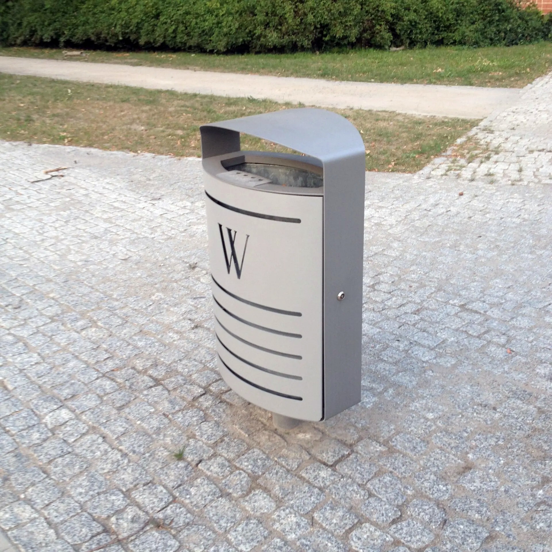 Szara metalowa kosz na śmieci z daszkiem i wyciętym motywem poziomych pasków oraz literą 'W', ustawiona na brukowanym chodniku w parku.