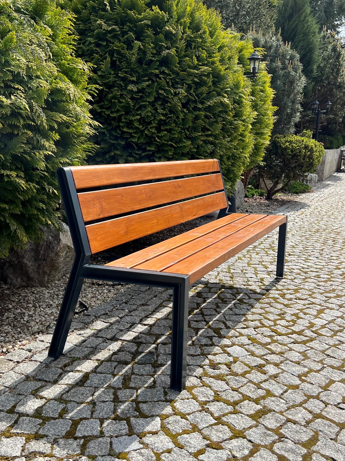 Nowoczesna ławka parkowa z drewna i czarnego metalu, ustawiona na brukowanej ścieżce w otoczeniu zielonych krzewów i drzew.