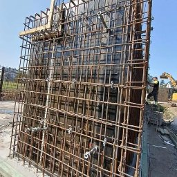 Płyty fundamentowe, wymiana gruntu, budowa domu - BUD-MAX - Szczegółowy widok szalunku zbrojeniowego z metalowymi ściągami na budowie w Krakowie, z pracownikiem i koparką w tle.