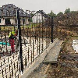 Płyty fundamentowe, wymiana gruntu, budowa domu - BUD-MAX - Zbrojenie pionowe na fundamencie betonowym, widoczny pracownik w odblaskowej kurtce, w tle domy jednorodzinne i wykop ziemny. Prace budowlane SSO w Krakowie.