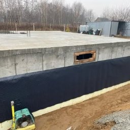 Płyty fundamentowe, wymiana gruntu, budowa domu - BUD-MAX - Betonowy fundament domu jednorodzinnego z podpiwniczeniem w trakcie izolacji, widoczna czarna folia kubełkowa i żółta izolacja termiczna oraz ubijarka wibracyjna.