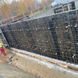Płyty fundamentowe, wymiana gruntu, budowa domu - BUD-MAX - Przygotowanie szalunku i zbrojenia pod wylewkę fundamentów domu jednorodzinnego z podpiwniczeniem, widoczne metalowe elementy mocujące i pracownik w odzieży roboczej.