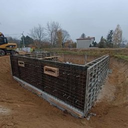 Płyty fundamentowe, wymiana gruntu, budowa domu - BUD-MAX - Szare szalunki zbrojeniowe w wykopie pod budowę domu jednorodzinnego z podpiwniczeniem, widoczny żółty dźwig w tle i pochmurne niebo.