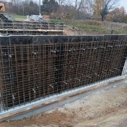 Płyty fundamentowe, wymiana gruntu, budowa domu - BUD-MAX - Zbrojenie pionowej ściany szalunkowej z widocznymi elementami mocującymi, przygotowane do wylania betonu, na tle wykopu i żółtej koparki w oddali.