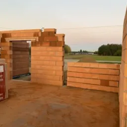 Wznoszenie ścian parteru domu jednorodzinnego z bloczków ceramicznych Porotherm, widoczne fragmenty ścian, otwór drzwiowy, paleta bloczków, w tle hałda ziemi i zabudowania.