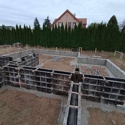 Płyty fundamentowe, wymiana gruntu, budowa domu - BUD-MAX - Przygotowanie pod fundamenty domu jednorodzinnego: szalunki ustawione na obrysie, widoczne zbrojenia i wykop, w tle rząd wysokich tuj oraz dom z czerwonym dachem.