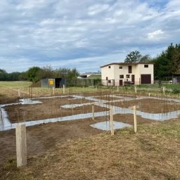 Płyty fundamentowe, wymiana gruntu, budowa domu - BUD-MAX - Fundamenty domu jednorodzinnego w Bogumiłowicach: zalane betonem ławy fundamentowe z wystającymi prętami zbrojeniowymi, widok na budynek gospodarczy i dom w tle.