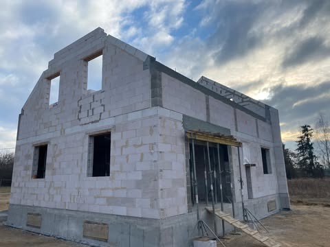 Surowy stan domu jednorodzinnego z białych bloczków, widoczne otwory okienne i drzwiowe, częściowo zadaszone wejście, w tle pochmurne niebo i drzewa.