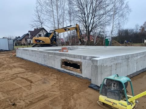 Wylany fundament z oknem piwnicznym w Tarnowie, widoczny żółty mini-koparka CAT oraz zagęszczarka gruntu AMMANN, przygotowany teren pod budowę domu.