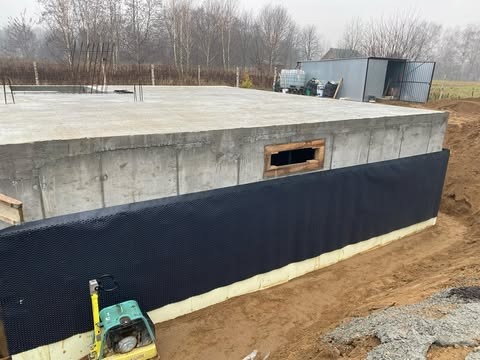Betonowy fundament domu jednorodzinnego z podpiwniczeniem w trakcie izolacji, widoczna czarna folia kubełkowa i żółta izolacja termiczna oraz ubijarka wibracyjna.